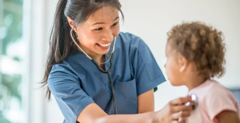 A smiling nurse practitioner listens to a young patient’s heart with a stethoscope.