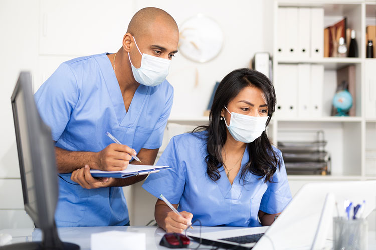 Two nurses wearing PPE masks take notes while consulting a computer screen.